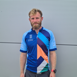 A person wearing a cycling top with blue sleeves and a colorful geometric pattern on the front, featuring the Stewart Brewing logo.