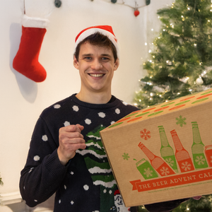 Man wearing a Christmas sweater and Santa hat holding a beer advent calendar in front of a decorated Christmas tree.