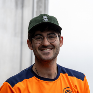 Man wearing a green cap and orange and navy shirt with a logo, standing against a plain background.