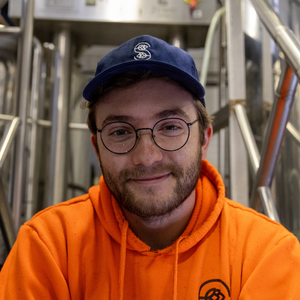 Person wearing an orange hoodie and blue cap with a logo, standing in front of industrial equipment.