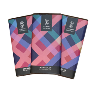 Three wrapped chocolate bars from Stewart Brewing are arranged in a fan shape on a plain white background. The wrappers feature a colorful geometric pattern with pink, blue, purple, and teal shapes, and black bands at the top and bottom with white text reading 'STEWART BREWING' and 'CRANACHAN WHITE CHOCOLATE'.