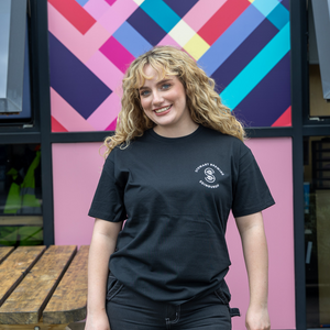 A person wearing a black crew neck t-shirt with a logo on the chest, standing in front of a colorful geometric patterned wall.